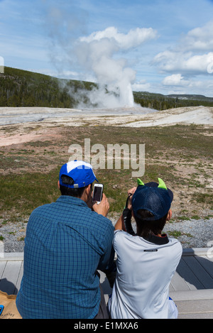 Un padre e figlio prendere immagini di telefono cellulare di geyser Old Faithful eruzione. Parco Nazionale di Yellowstone, Wyoming negli Stati Uniti. Foto Stock