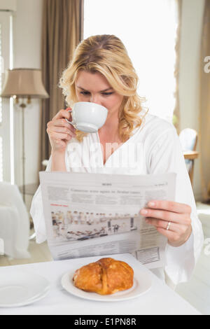 Bella bionda in accappatoio a bere caffè e quotidiano di lettura Foto Stock