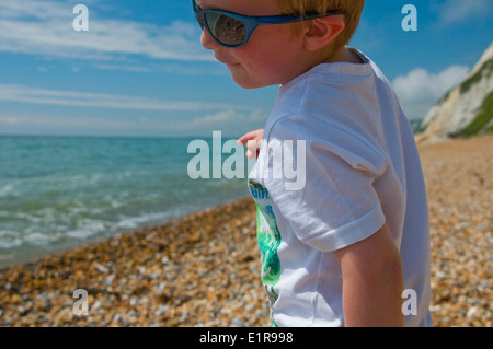 Close up di un giovane ragazzo giocando su una spiaggia di ciottoli con occhiali da sole Foto Stock