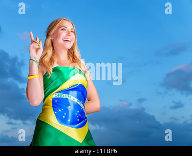 Felice fan del calcio brasiliano squadra, allegro Pretty girl su stadium il tifo a sostegno, indossando grande bandiera nazionale del Brasile Foto Stock