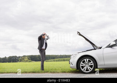 Triste giovane imprenditore in piedi con le mani sulla testa in prossimità broken auto a campagna Foto Stock