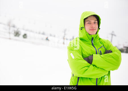 Ritratto di giovane uomo in giacca con cappuccio in piedi con le braccia incrociate nella neve Foto Stock