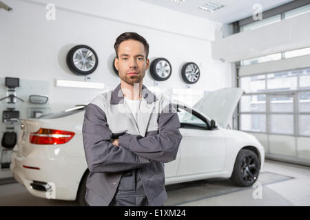 Ritratto di fiducioso maschio meccanico di automobili in piedi nella parte anteriore della vettura in officina Foto Stock