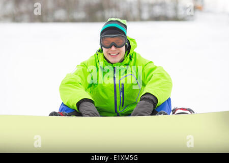 Ritratto di giovane sorridente uomo con lo snowboard seduta sul terreno nevoso Foto Stock