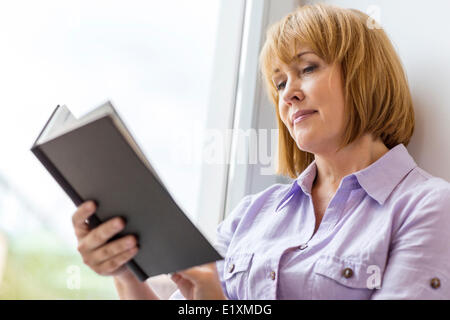 Donna matura libro dalla finestra a casa Foto Stock