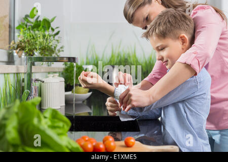 Madre che assiste il figlio in manicotti di piegatura durante il lavaggio delle mani in cucina Foto Stock