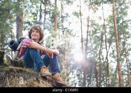 Per tutta la lunghezza del maschio sorridente escursionista che guarda lontano mentre è seduto sulla scogliera in foresta Foto Stock