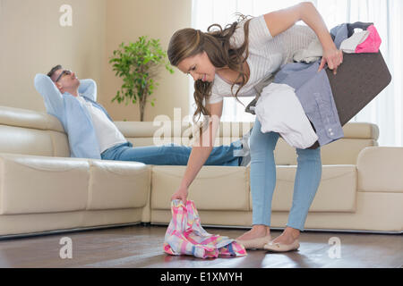 Donna con cesto per la biancheria di prelievo mentre abbigliamento uomo rilassante sul divano in background Foto Stock