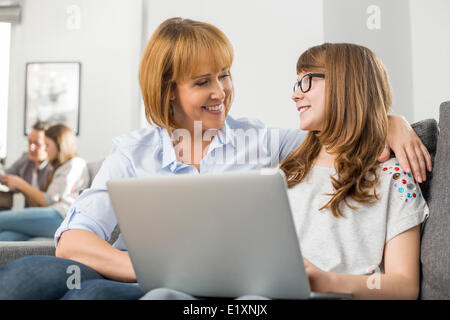 Felice madre e figlia con il computer portatile mentre la famiglia seduta in background a casa Foto Stock