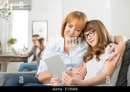 Affettuosa madre e figlia con tavoletta digitale con la famiglia seduta in background a casa Foto Stock