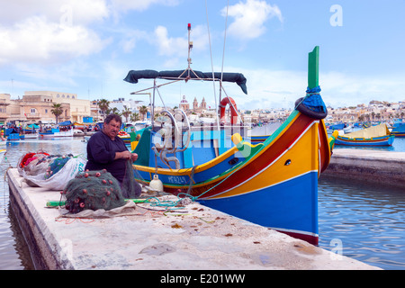 Fisherman sat sul quay riparare reti accanto alla sua barca nel porto di Marsaxlokk, Malta, Foto Stock