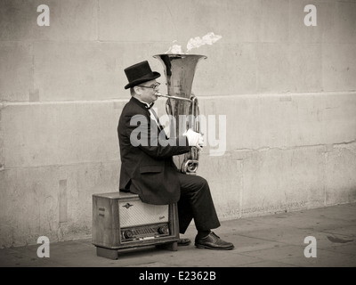 Busker, Christopher Werkowicz giocando flaming tuba seduto su una vecchia radio contro un semplice muro di pietra in seppia Foto Stock