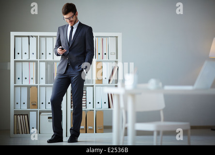 Bello imprenditore della tuta e occhiali la scrittura di sms in ufficio Foto Stock