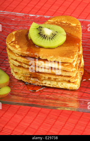 Pila di heart-shaped pancake con sciroppo e kiwi su un vetro trasparente piatto su uno sfondo rosso. Foto Stock