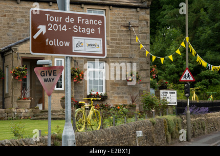 Brown tourist road sign, decorativi bunting & bici gialla sul display in giardino - prima Le Tour de France, villaggio Addingham, nello Yorkshire, Inghilterra, Regno Unito. Foto Stock