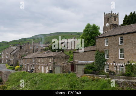 Tradizionale cottage in pietra il rivestimento stradale, chiesa torre & colline oltre - Muker, piccolo borgo rurale in Swaledale, Yorkshire Dales, Inghilterra, Regno Unito. Foto Stock