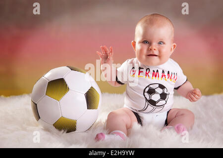 Baby girl, 7 mesi, indossando una Germania football jersey con un calcio, colori tedesco sul retro Foto Stock