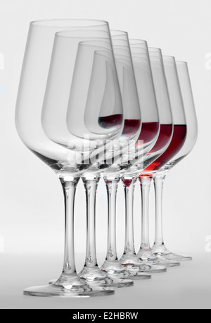 Alcuni bicchieri di vino in piedi consecutivi in una fila in grigio chiaro indietro, uno parzialmente riempito con un bicchiere di vino rosso Foto Stock