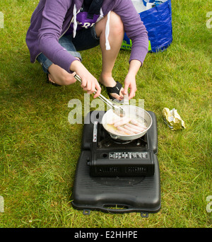 Una giovane donna in flip flop cuochi salsicce per la colazione su un fornello da campeggio. Foto Stock