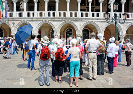 Gruppo di turisti nel tour di Venezia in piedi e ascoltando la guida (alzata a mano) fuori dal museo del Palazzo Ducale in una giornata di sole molto caldo così il sole cappello Foto Stock