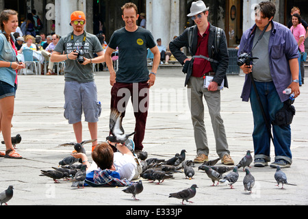 Persone spettatori che chiacchierano con la donna che si stende su lastre di pavimentazione giorno caldo in Piazza San Marco Venezia dando da mangiare ai piccioni dalle sue mani Italia Europa Foto Stock