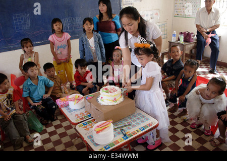 Un insegnante è aiutare un bambino di 6 anni ragazza asiatica tagliare la sua torta di compleanno presso la Scuola di Angkor in Kampong Cham, Cambogia. Foto Stock