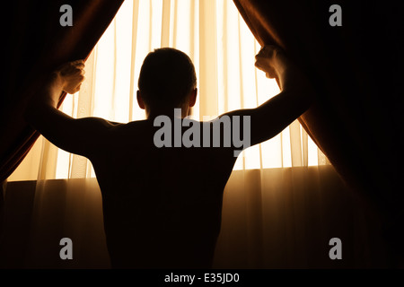 Uomo in sala buia apre tende su finestra per la luce del mattino Foto Stock