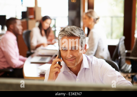 Imprenditore sul telefono alla scrivania con sale riunioni in background Foto Stock