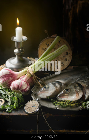 Natura morta con pesce crudo di spigola, erbe aromatiche, ortaggi e orologio vintage Foto Stock