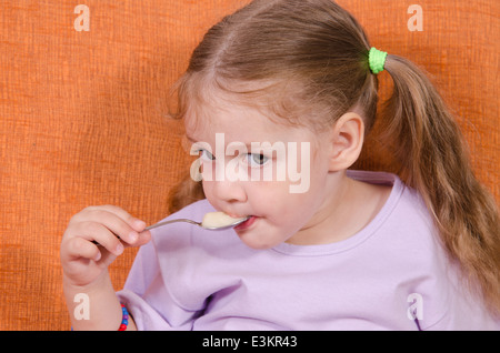 Bambina di tre anni con piacere e umorismo mangia miele cucchiaio seduta sul lettino Foto Stock