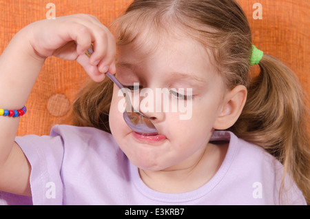 Bambina di tre anni con piacere e umorismo mangia miele cucchiaio seduta sul lettino Foto Stock