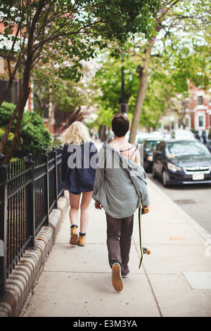 Giovane uomo adulto che trasportano skateboard seguenti giovane donna camminando lungo la strada Foto Stock