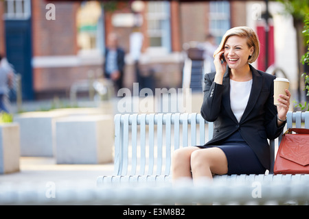 Imprenditrice su una panchina nel parco con caffè utilizzando il telefono cellulare Foto Stock