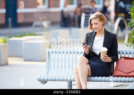 Imprenditrice su una panchina nel parco con caffè utilizzando il telefono cellulare Foto Stock