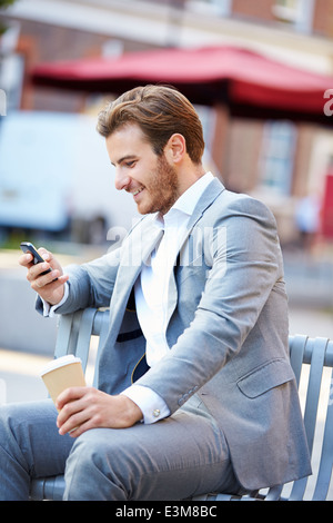 Imprenditore su una panchina nel parco con caffè utilizzando il telefono cellulare Foto Stock