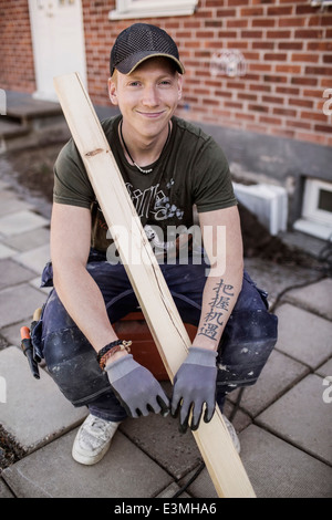 Ritratto di falegname sorridente con asse in legno seduta al sito in costruzione Foto Stock