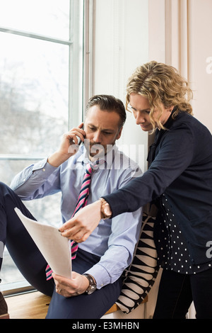 Imprenditore rispondendo alle smart phone mentre discutete su documento con il collega femmina dalla finestra di ufficio Foto Stock