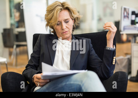 Imprenditrice matura la lettura di documenti mentre si è seduti su una sedia in ufficio Foto Stock