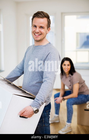 Ritratto di uomo felice che porta una scatola di cartone con la donna in background a casa Foto Stock