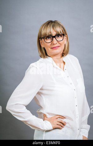 Imparato attraente di mezza età donna bionda in bicchieri in piedi guardando la telecamera con bracci ripiegati contro un backgroun grigio Foto Stock