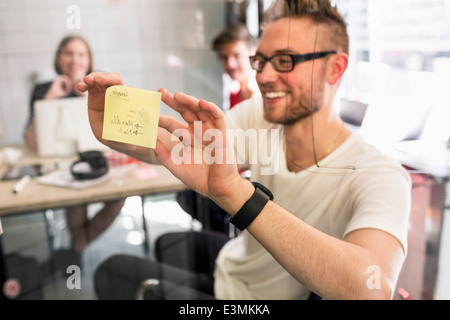 Giovane imprenditore incollaggio nota adesiva sul vetro trasparente con i colleghi a sfondo nel nuovo ufficio Foto Stock