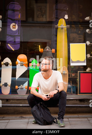 Per tutta la lunghezza della giovane uomo seduto al di fuori del negozio di skateboard Foto Stock