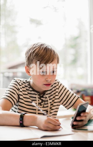 Boy utilizzando smart phone durante la scrittura a tavola in casa Foto Stock
