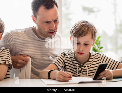 Boy utilizzando il telefono cellulare mentre la scrittura a tavola da padre in casa Foto Stock