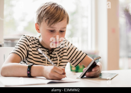 Boy utilizzando il telefono cellulare durante la scrittura in un libro a tavola Foto Stock