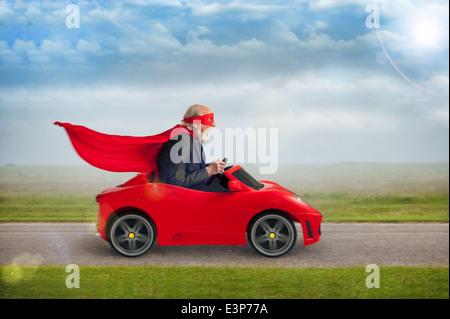 Il supereroe senior uomo con maschera e la guida del capo di un giocattolo auto sportiva rossa Foto Stock