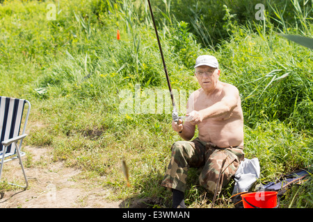 Anziani disabili pescatore seduto su una riva del lago la bobinatura in un piccolo pesce sulla sua asta e la linea Foto Stock