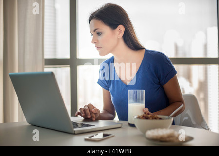 Giovane donna utilizzando laptop mentre si consuma la colazione Foto Stock