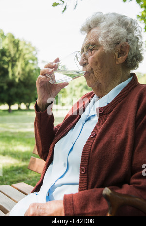 Senior donna seduta su una panchina nel parco, bere un bicchiere di acqua Foto Stock