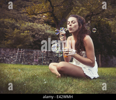 Giovane donna seduta nel campo a soffiare bolle Foto Stock